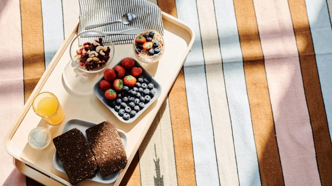 Top view of a healthy breakfast tray with fruits, bread, and juice on a striped tablecloth.