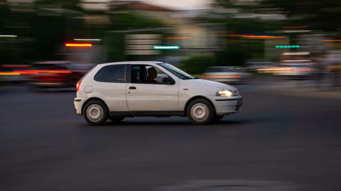 A white car speeds through a city street in the evening, showcasing motion blur.